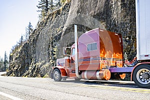 Extended cab orange big rig classic semi truck transporting cargo in dry van semi trailer driving on the mountain road with rocks