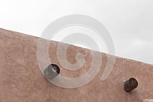 Exterior of red adobe building, two exposed vigas, grey cloudy skies