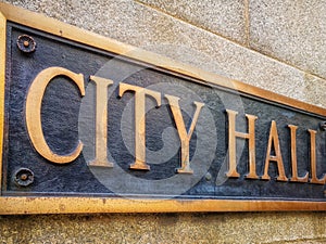 Exterior sign that reads `City Hall ` in brass lettering. Close up