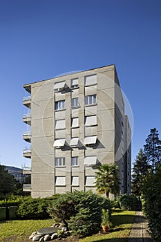 Exterior view of an old rough concrete apartment building with balconies and blinds. There is a green garden and trees around