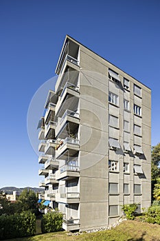 Exterior view of an old rough concrete apartment building with balconies and blinds. There is a green garden and trees around