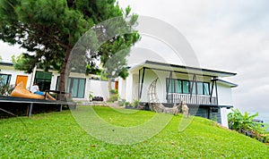 Exterior view of white house on hill with tree and green grass lawn. Woman relaxing under the tree outside home. Modern