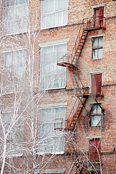 External fire escape staircase on an old brick building of factory, plant. Big windows, white birch trees grows near.