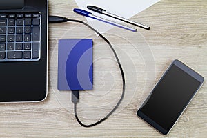 External hard drive connecting to laptop with smartphone, pens and paper on wooden desk