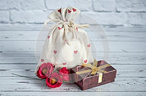 Extile bag with a gift. Red hearts are depicted on the fabric. Nearby lies a box with a gold bow and three scarlet flowers. Close-