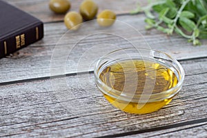 Extra virgin olive oil in a glass container with a closed Holy Bible Book, green olive branch, and olives on a wooden table