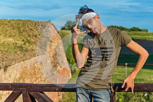 Extravagant hipster male model with sunglasses and a white scarf on a wooden bridge. The model shows the finger as a devilish si