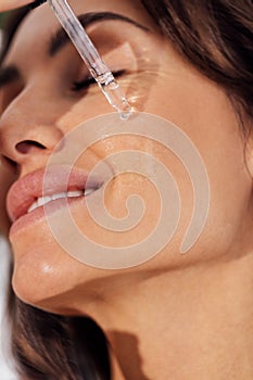 Extreme close up of beautiful smiling caucasian girl. Young woman applies transparent serum from pipette to her face