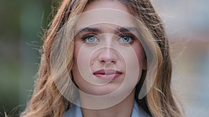 Extreme close-up female serious face with natural makeup blue eyes looking at camera posing outdoors. Young Caucasian