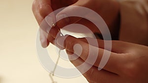 Extreme close-up hands of unrecognizable woman putting thread through eye of needle on blurred light background.