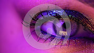 Extreme close up of human eye iris under violet neon light. Female with beautiful makeup, glitter shadows. Womens green