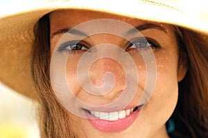 Extreme close up of young model face with straw hat smiling at camera under summer rays sun on the beach.