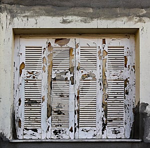 Extremely chipped and weather-worn white paint on closed grungy wooden shutters in stucco building with grey paint smeared abound photo