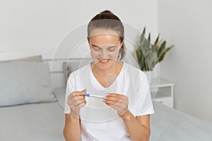 Extremely happy attractive woman wearing casual attire sitting on sofa at home and looking at pregnancy test with positive result