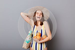 Extremely happy caucasian woman wearing striped swimsuit and straw hat  gray background holding oranges in hands rejoicing