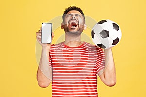 Extremely happy excited man with beard in striped t-shirt sincerely laughing holding white display smartphone and soccer ball,