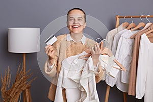 Extremely happy joyful woman with bun hairstyle wearing beige jumper standing against gray wall with clothes on hangers on shelf,