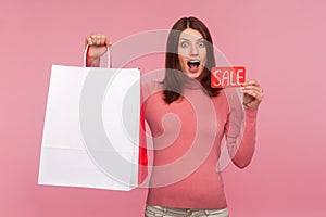 Extremely happy surprised woman holding in hands paper shopping bags and sale card, looking at cameras with big eyes and opened