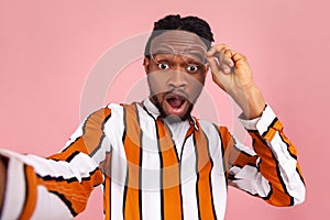 Extremely shocked surprised african man blogger with dreadlocks in striped shirt putting off eyeglasses and looking at selfie