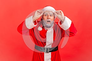 Extremely shocked surprised santa claus looking at camera with big wondered eyes holding eyeglasses on forehead, holidays coming