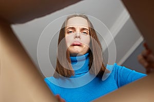 Extremely upset woman customer wearing blue turtleneck opening carton box, looking inside parcel with pout lips, crying,