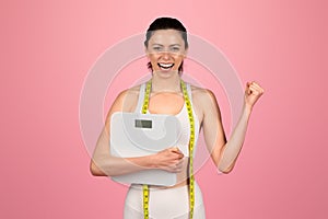 Exultant young woman celebrating her weight loss success, holding a scale and wearing a measuring tape