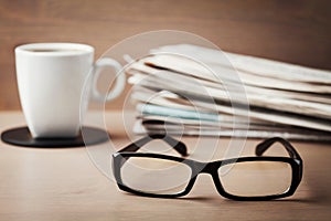Eyeglasses, coffee mug and stack of newspapers on wooden desk for themes of ophthalmology, poor vision and reading photo