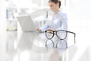 Eyeglasses leaning on desk and woman working on computer at offi