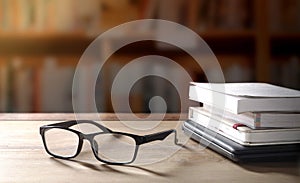 Eyeglasses with stacked books and black laptop on wooden table in library