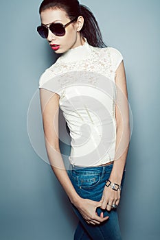 Eyewear concept. Fashionable beautiful young woman in trendy clothing white waistcoat, blue jeans, sunglasses over blue