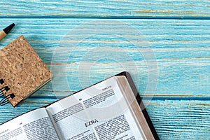 Ezra open Holy Bible Book with a small notebook and pen on a rustic wooden background