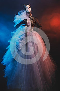 Fabulous creature, queen, sorceress, in a colorful suit and sword. Woman, model after 60, in the studio, dark background