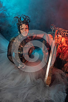 Fabulous creature, queen, sorceress, in a colorful suit and sword. Woman, model after 60, in the studio, dark background