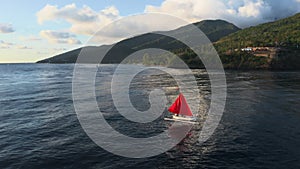 Fabulous scarlet red sail boat floating on quiet ocean water near sea shore with green hills and mountains in summer