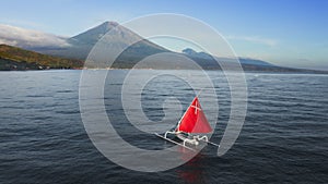 Fabulous scarlet red sail white Jukung boat floating on quiet ocean water near sea shore with huge volcano in summer