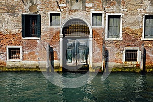 Facade of partially mossy old brick house with wooden vintage door on narrow canal in Venice, Italy.