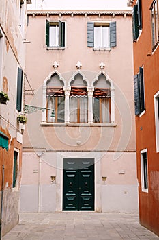 Facade of a three-story peach house in Venice, Italy. Massive green wooden front door, three classic Venetian windows