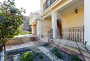 The facade of a two-story white classic cottage with a front garden. Carved entrance door, car gate, beige tiles.