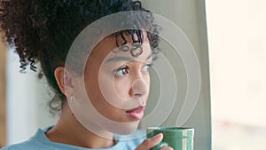 Face of beautiful african woman blowing her hot coffee or tea while looking out the window and relaxing at home. Serious