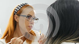 Face of beautiful red-haired makeup artist in glasses does makeup to a young woman in beauty salon