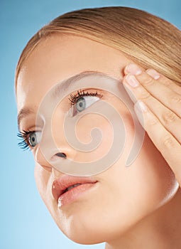 Face of beautiful woman, cosmetic skincare on blue background in studio and side profile with hand on temple. Young