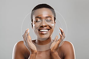 Beautiful afro woman applying cream on her cheeks and sincerely smiling photo