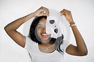 Face care and beauty treatments. Pretty smiling African woman holding a sheet moisturizing mask near her face, standing