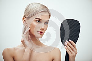 Face care. Portrait of gorgeous blonde woman touching her face and looking in small mirror while standing against grey
