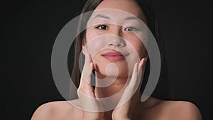 Face care procedures. Young beautiful asian lady caressing her face, enjoying her smooth skin, black background