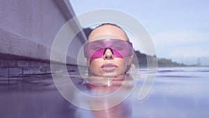 Face closeup of young woman wearing purple sunglasses looking at camera in infinity rooftop swimming pool on a sunny day.