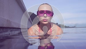 Face closeup of young woman wearing purple sunglasses looking at camera in infinity rooftop swimming pool on a sunny day.