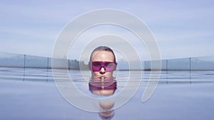 Face closeup of young woman wearing purple sunglasses looking at camera in infinity rooftop swimming pool on a sunny day.