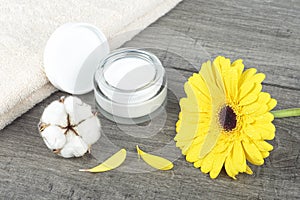 Face cream jar on wooden background with yellow gerbera, soft face towel and cotton flower
