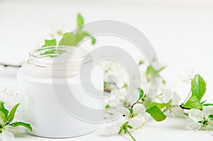 Face cream in white jar on a white background with white small flowers of an apple tree. Concept natural cosmetics